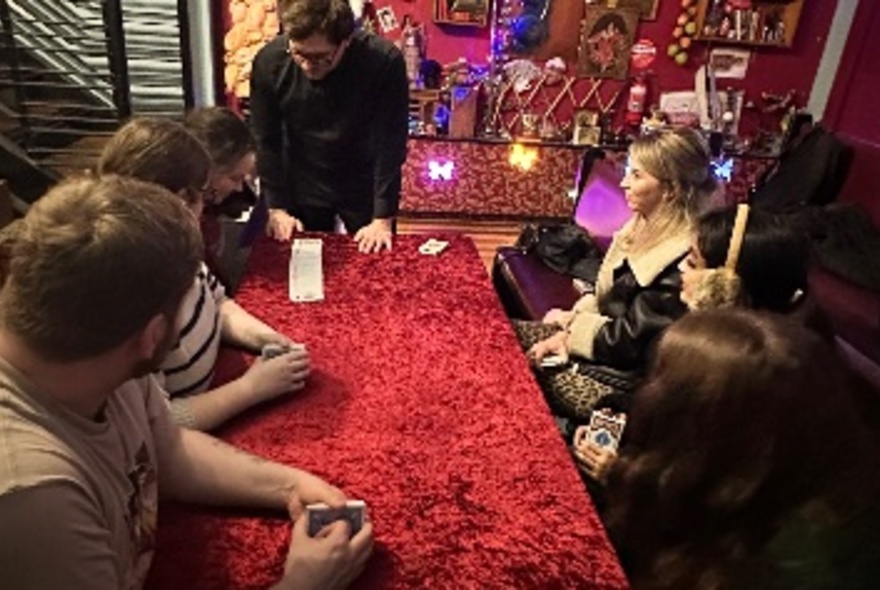 A group of people seated around a red velvet covered table learning how to do card tricks. 