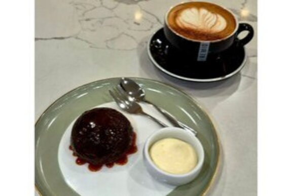 A chocolate pudding with raspberry sauce and cream in a small pot, alongside a cappucino in a mug.