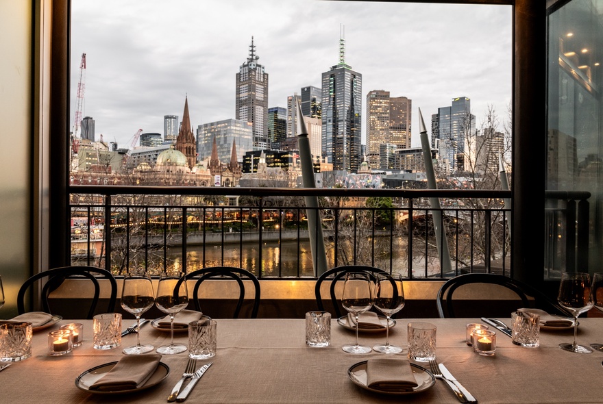 A long table in a restaurant set for dinner service, with a view out of the window behind the table of the Melbourne city skyline and the Yarra River.
