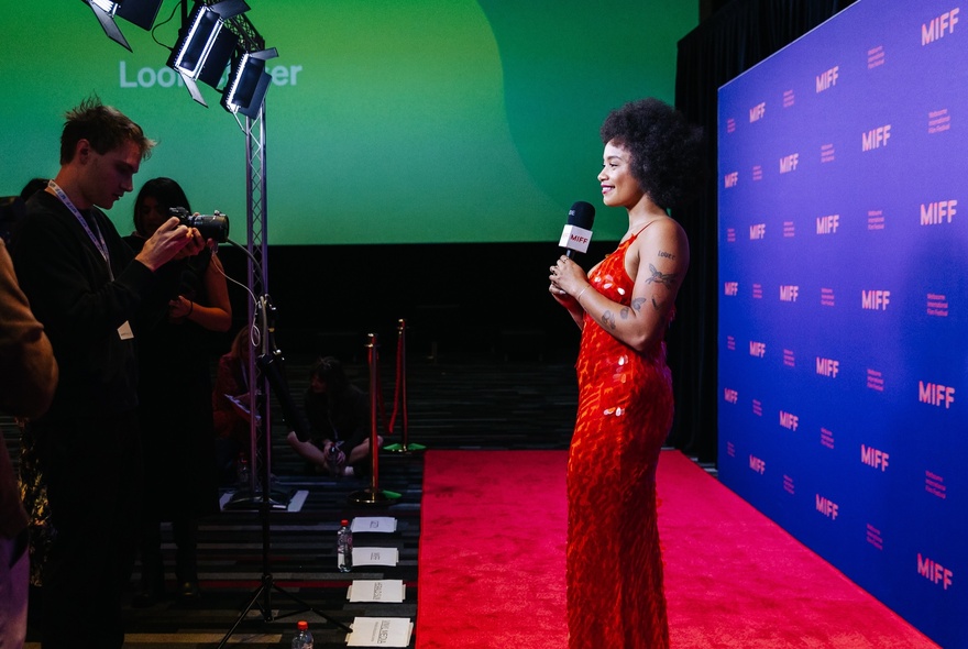 A red carpet premiere at the Melbourne International Film Festival, a person in a red dress holds a microphone, and talks to a group of photographers and people who stand in front of her.