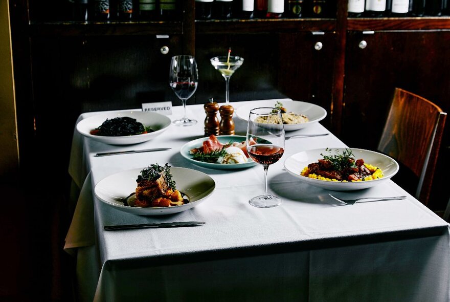 Restaurant table set for four with main courses, shared salad, glasses of wine, white napery.