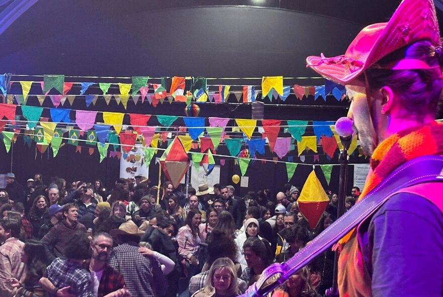 A crowded hall filled with people dancing underneath colourful bunting; in the right foreground a man is playing guitar.