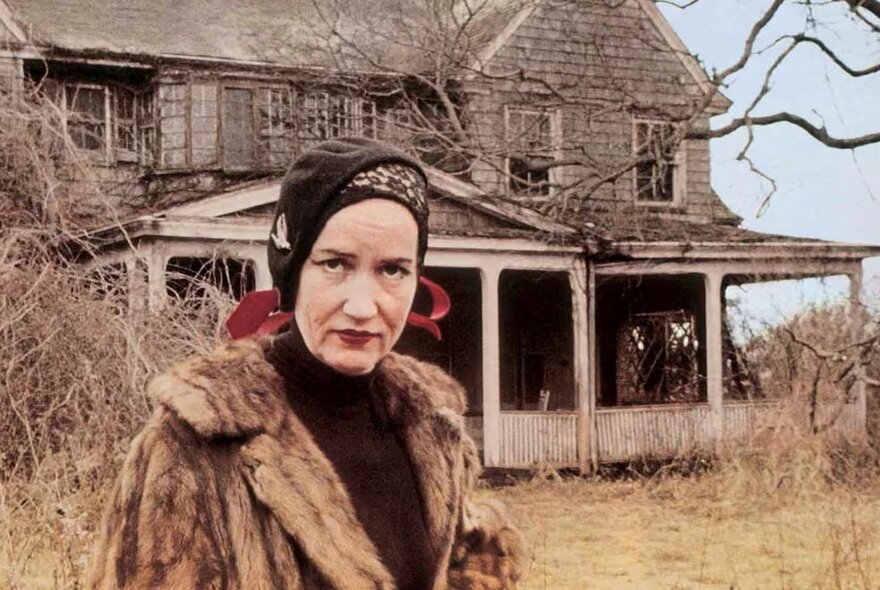 A colour photograph of Edith 'Little Edie' Bouvier Beale standing outdoors in front of her dilapidated Grey Gardens estate. She wears a black headscarf, a matching black turtleneck, and a large brown fur coat. 