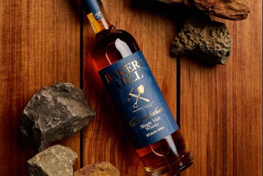 A bottle of Bakery Hill Single Malt Whisky lying flat on a timber table top next to small rocks.