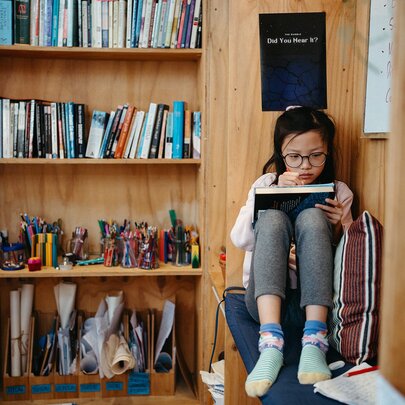A girl reading in a study nook next to a bookcase in a studio space, taking part in a creative writing workshop.