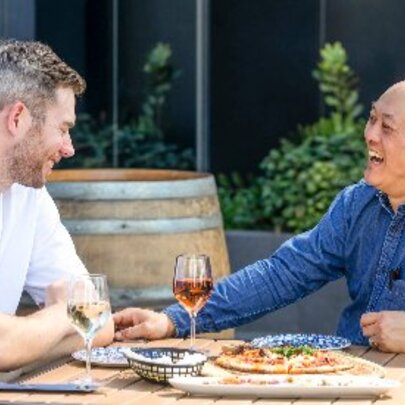 Two men enjoying pizza and wine at an outdoor table at a restaurant.
