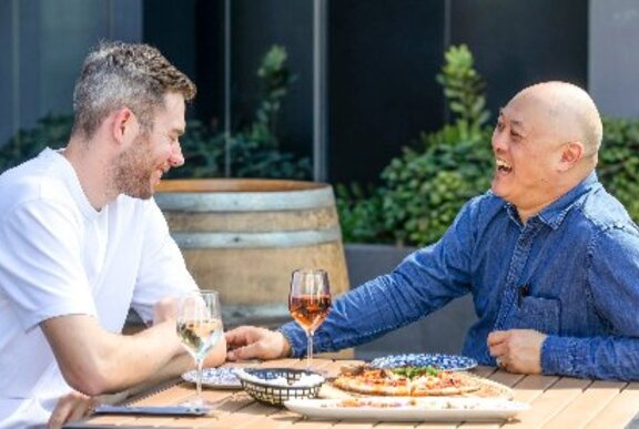 Two men enjoying pizza and wine at an outdoor table at a restaurant.