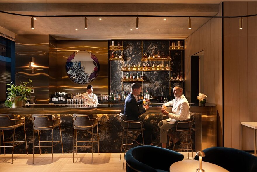 Two people seated on bar stools and drinking at an elegant bar, while a person behind the bar prepares drinks.