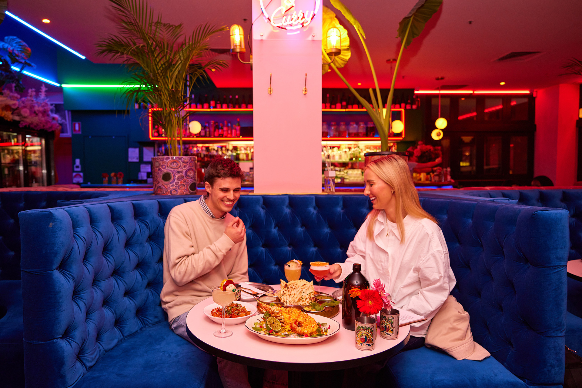 Two friends are seated in a booth at a brightly coloured restaurant eating Indian food and drinking cocktails