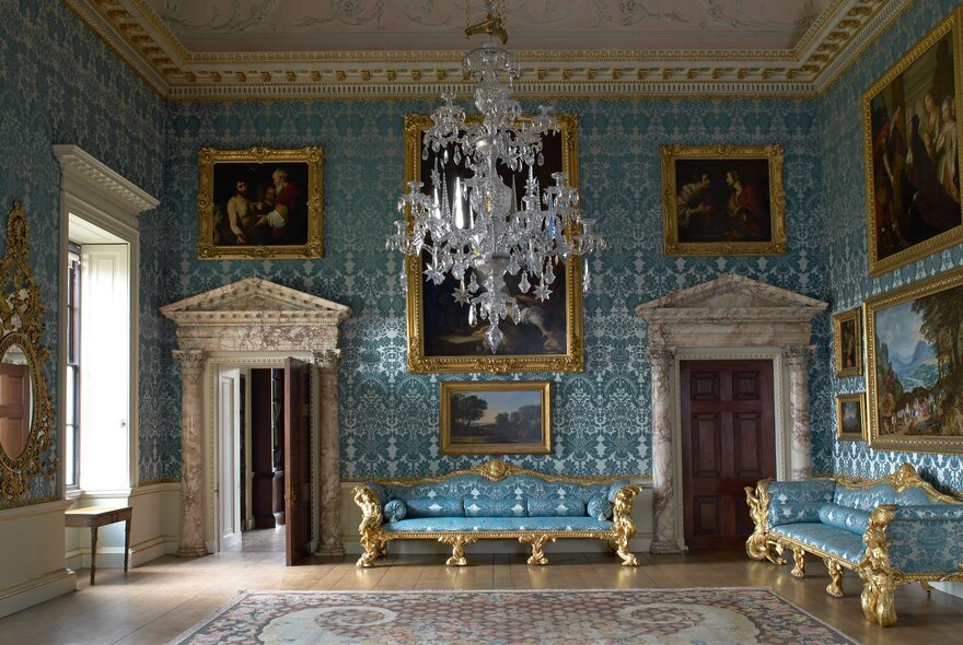 A blue room in an ornate British country house with a huge chandelier in the centre. 