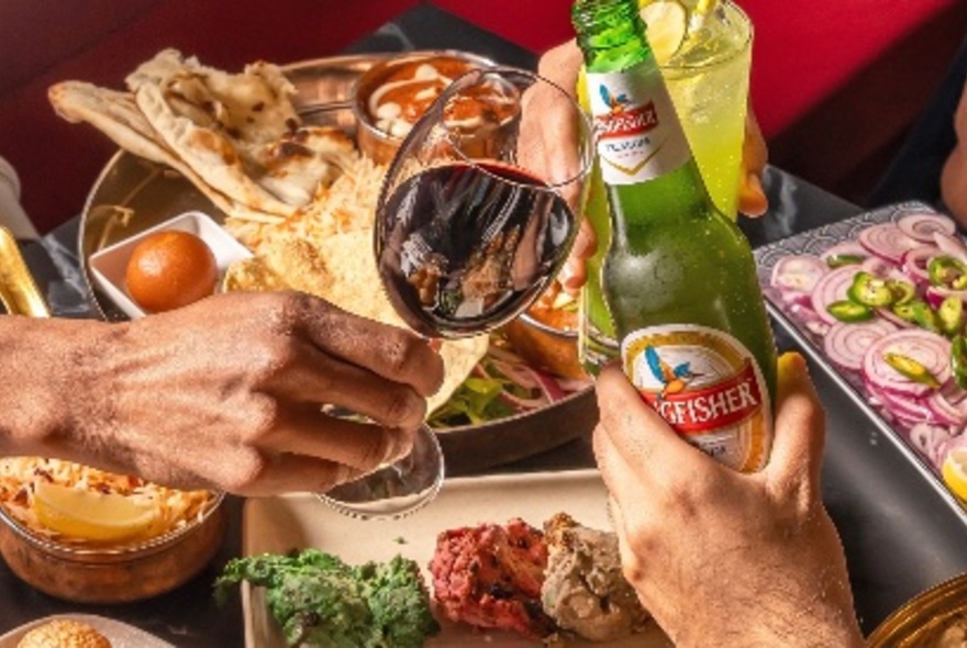 Two people's hands clinking drinks in front of a delicious array of North Indian dishes.