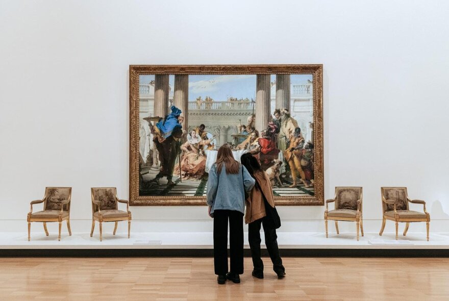 The back of two people gazing a a large painting in a gallery with two chairs either side of the painting. 