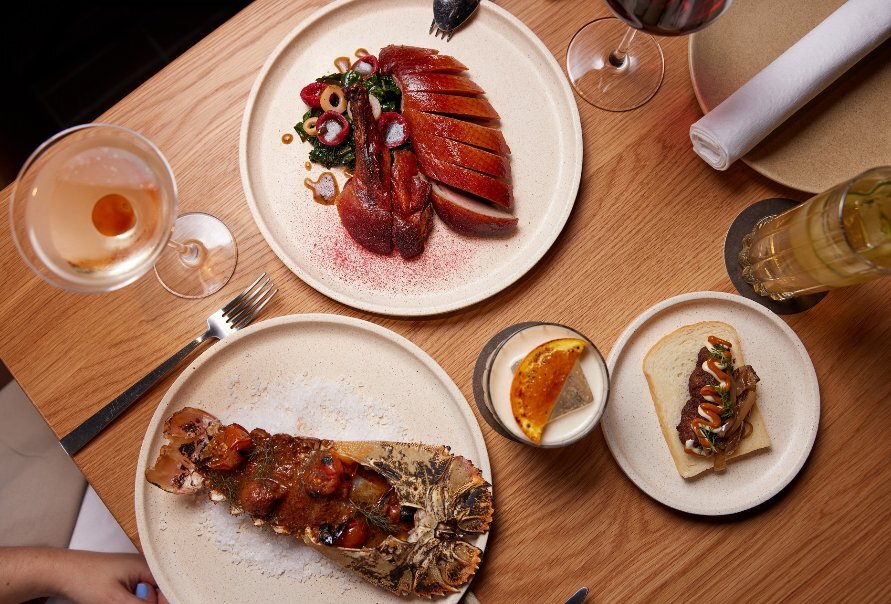 Wooden table with white plates of food including duck, lobster tail and cocktails.