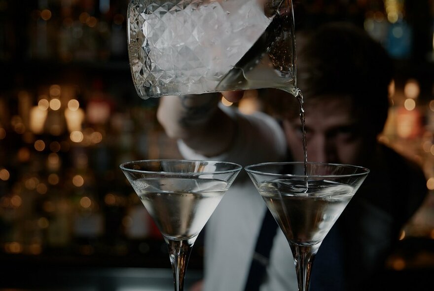 Bartender pouring martinis into two martini glasses in front of him.