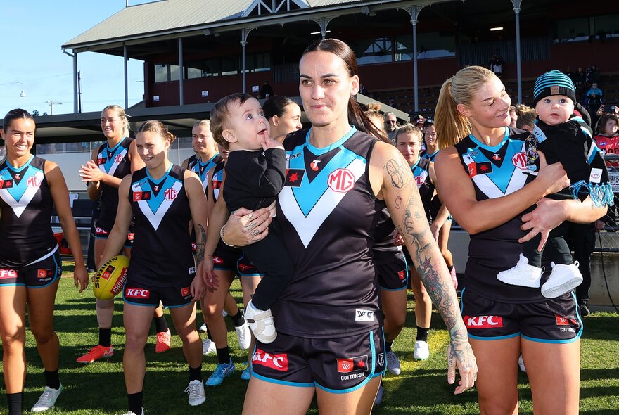 Port Adelaide AFLW players walking out onto the football field, some of them holding their babies and toddlers in their arms.