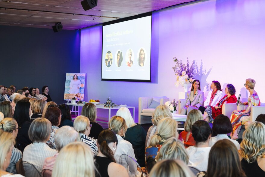 Inside at a seminar with a panel of women up front next to a large video screen and an audience looking on. 