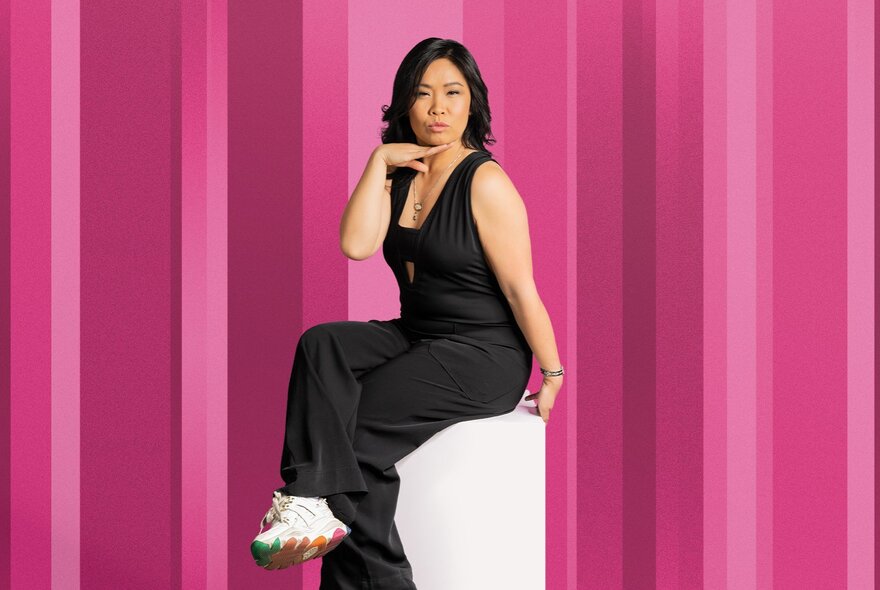 Comedian Diana Nguyen sitting on a white pedestal, posing, against a pink striped background. 