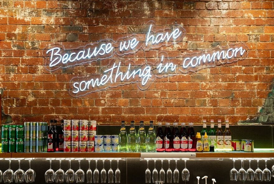 Brick wall with neon sign above lines of cans, bottles and glasses.