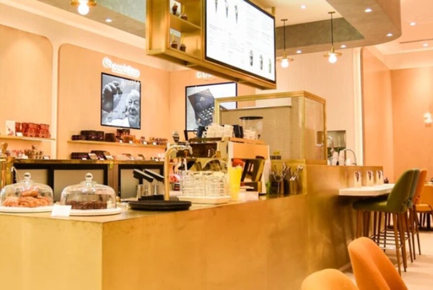 Godiva cafe interior with wooden counter with cakes under glass domes, tables and chairs, central overhead display.