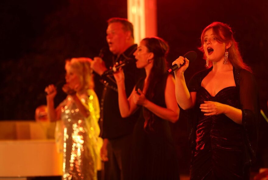 Four singers on stage, three in black and one in gold sequins, all holding microphones and singing.