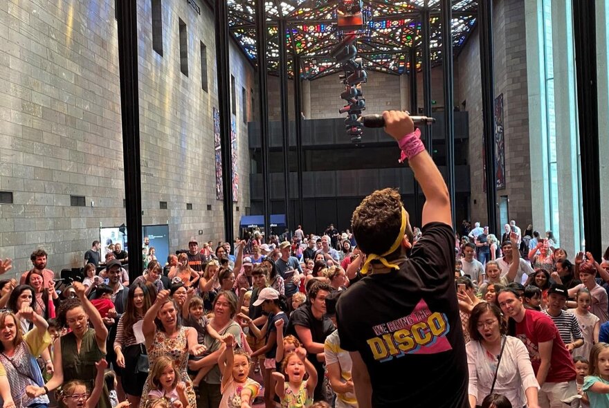 A crowd of adults and children dancing with a dance leader and DJ standing in front of them, in the great Hall at the NGV Kids Summer Festival.