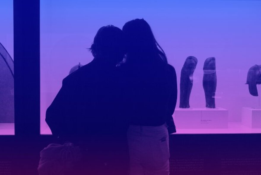 The back of two people cuddled together in front of an exhibition, the image has a pink and blue filter over it. 