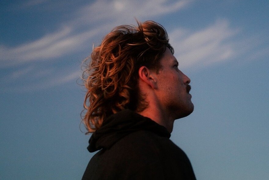 Musician, Ziggy Alberts, wearing a black hooded jumper and looking up at the blue sky with soft white clouds.