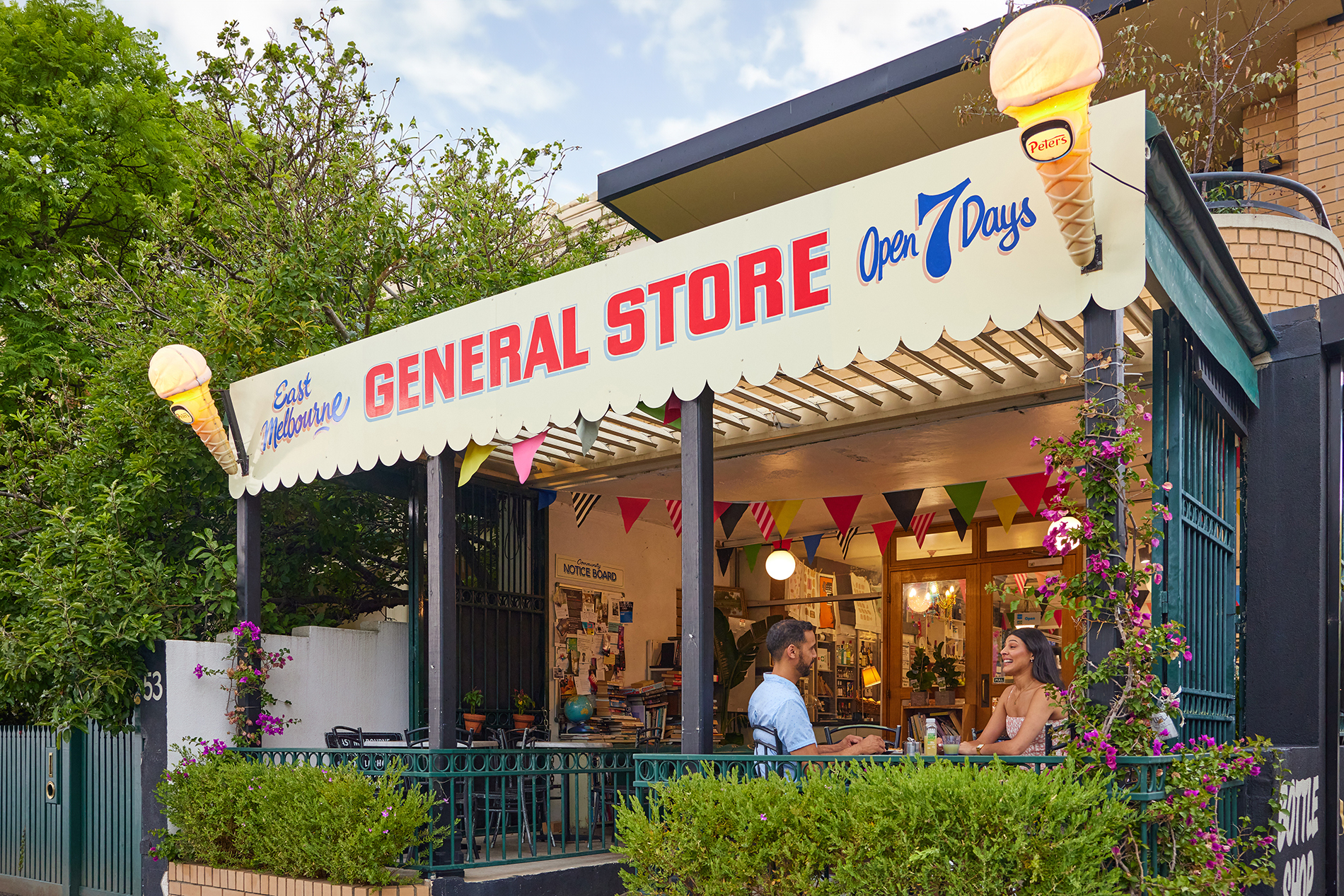Two people sitting outside a general store with bunting and a sign with ice creams.