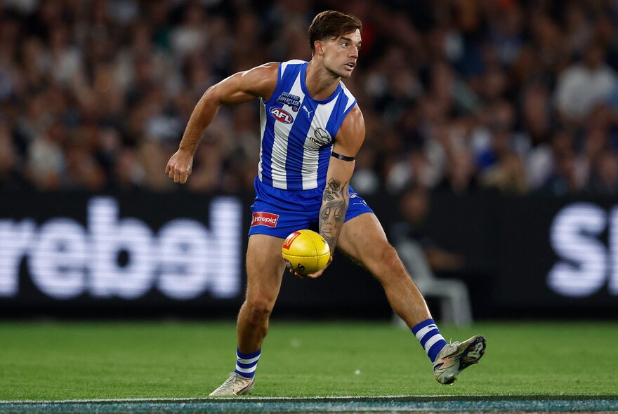 A North Melbourne AFL player about to handball a yellow football, a blurred crowd in the background.