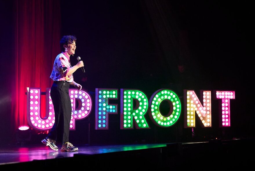 A comedian standing on a stage talking into a microphone, behind her is a sign spelling out the words UPFRONT in colourful lights.