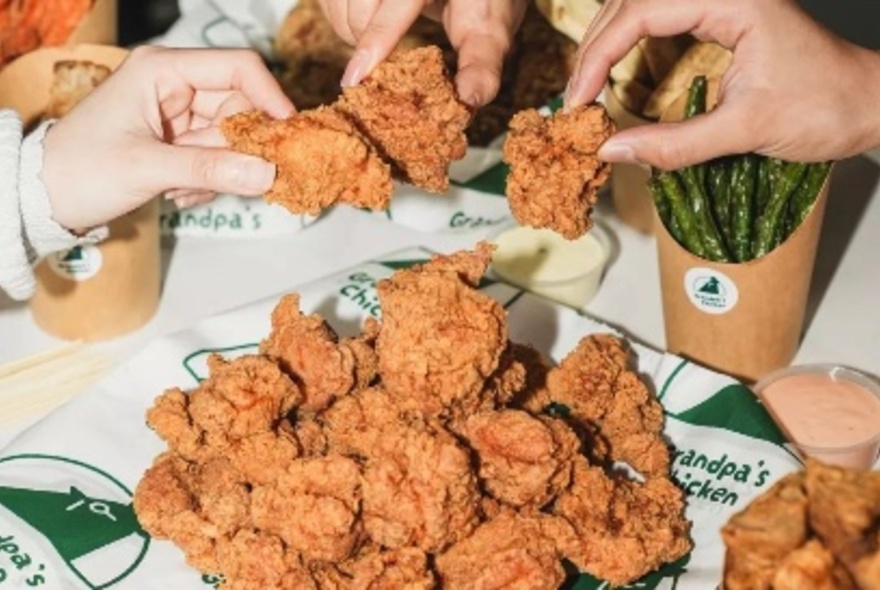 Hands holding fried chicken pieces over a platter of food.