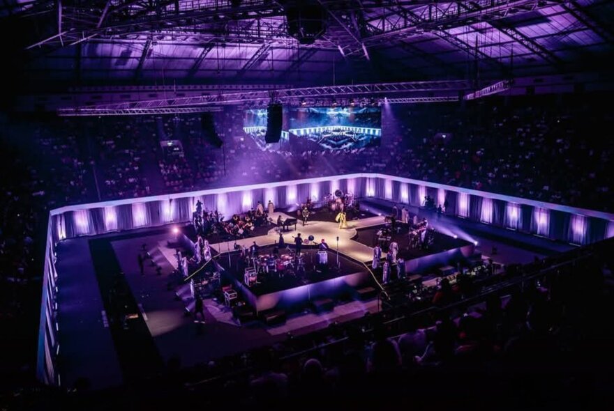 A music concert featuring a large group of performers on a brightly lit raised central stage, surrounded by an audience, in roofed arena.