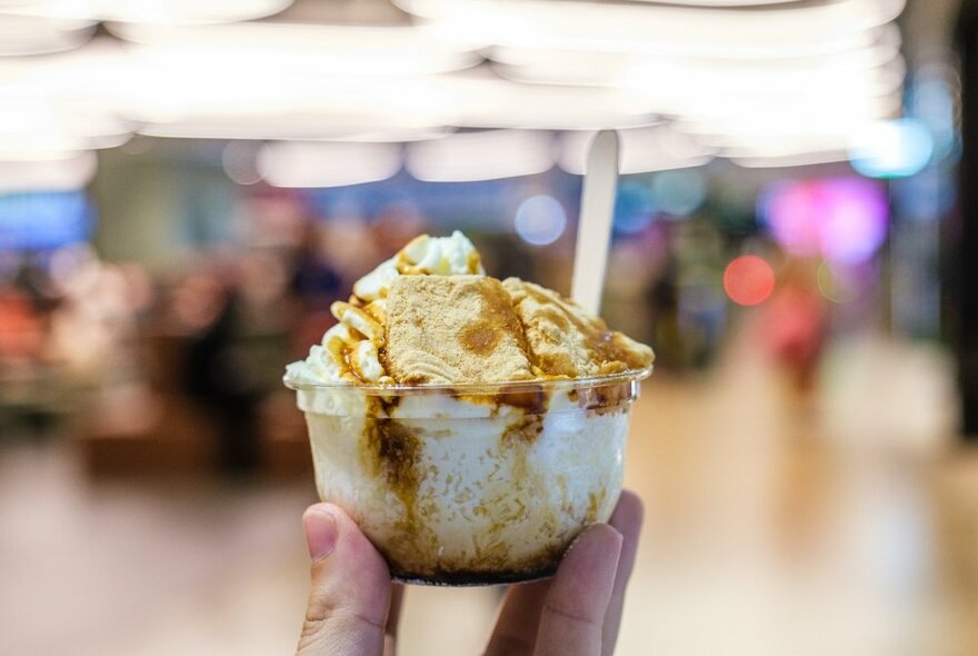 A hand holding up a clear plastic takeaway container of ice cream with a wooden spoon sticking out of it.