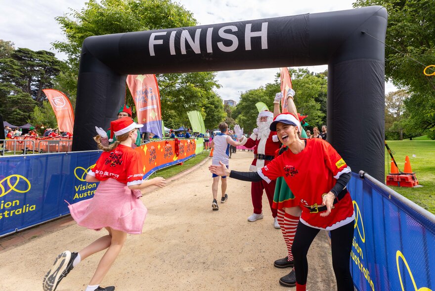 A runner wearing a Santa hat and pink dress passing under the finish line with Santa's elves and Santa.