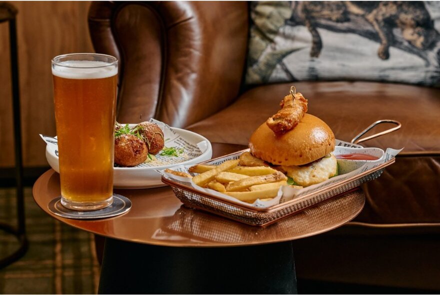 A small coffee table at a restaurant, laden with pub-fare including a burger and chips, arancini balls, and a glass of beer on a coaster.