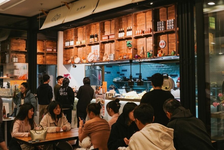 Diners at tables and ordering counter in a busy Asian cafe with wooden shelving details.