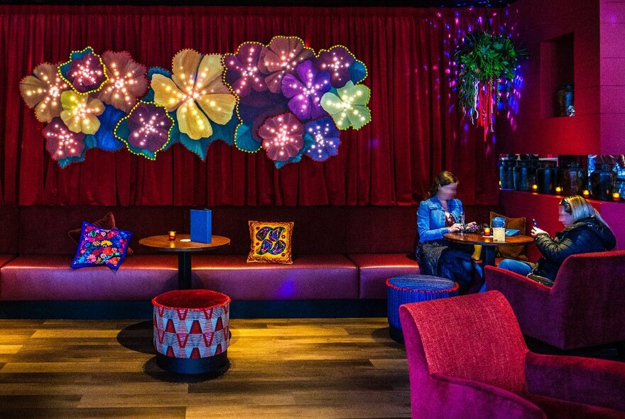 Two women dining in a restaurant with couches and a big flower art piece on a red curtained wall.