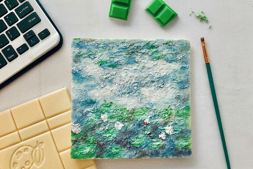 Square painted with green landscape, green paintbrush to right, loose green chocolate squares above, a keyboard to the right.