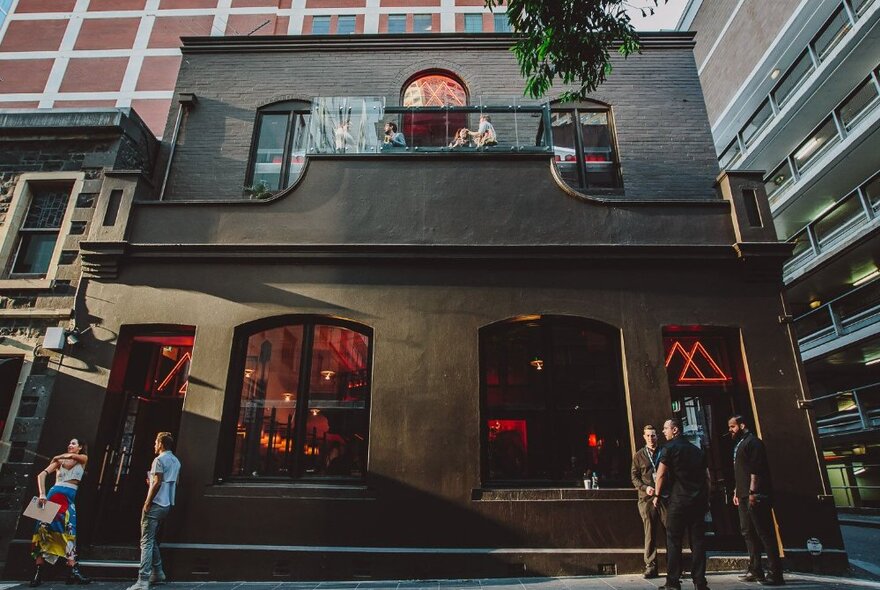 The dark grey shopfront of Magic Mountain Saloon with people standing out the front and red lighting in the windows.