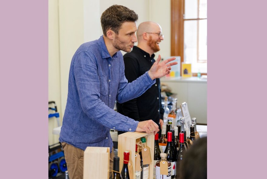 Two men talking in front of a table with bottles of wine.