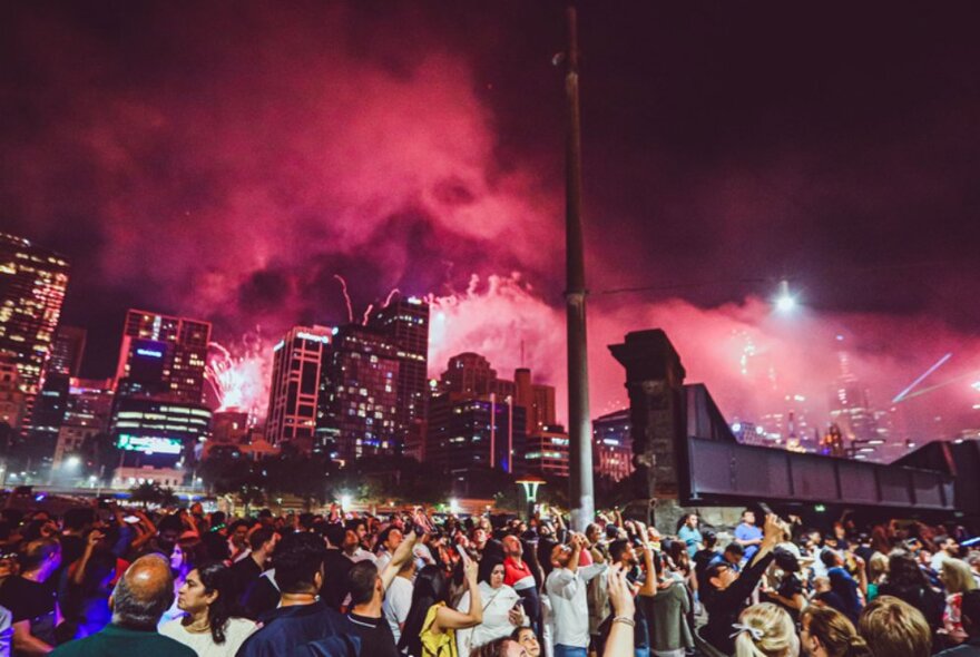 Red coloured fireworks going off over the Melbourne city skyline with a crowd of people visible on the streets. 