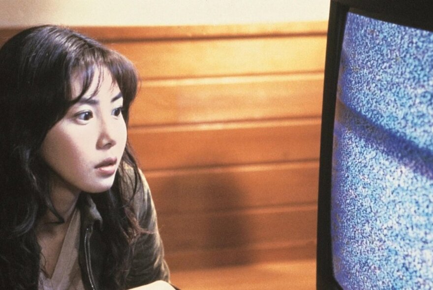 A still from a J-Horror movie of a young Japanese woman staring at a television with nothing but static on the screen.