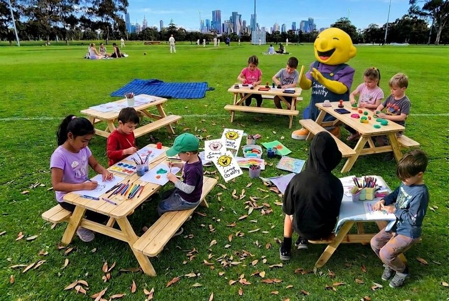 Small wooden picnic tables in a large open park, with kids seated at them taking part in craft activities. 