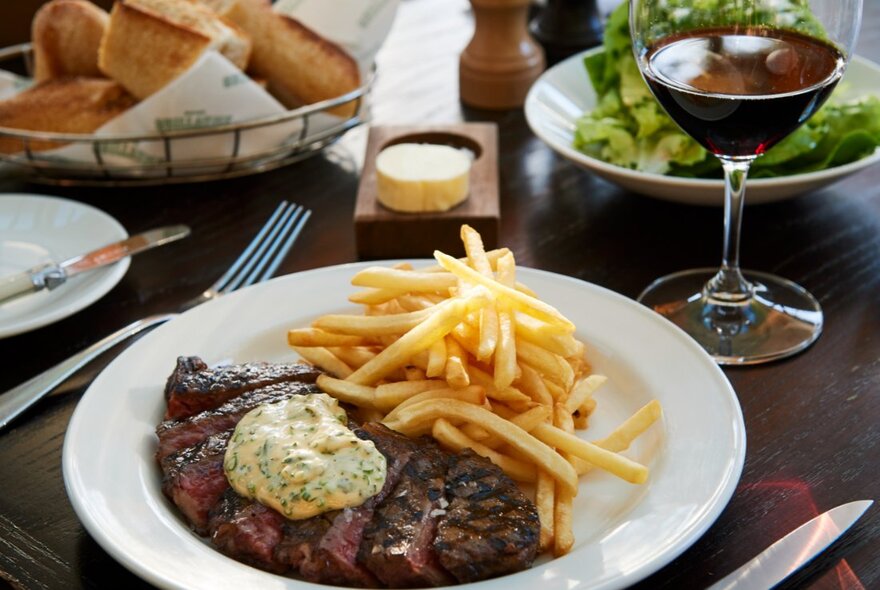 Steak frites served on a white plate, with a leafy green side salad, and a glass of red wine, on a restaurant dining table.