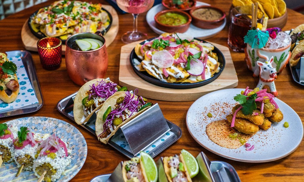 An assortment of colourful Mexican dishes spread out across a wooden table, including tacos, jalapeno poppers, nachos, cocktails and more.