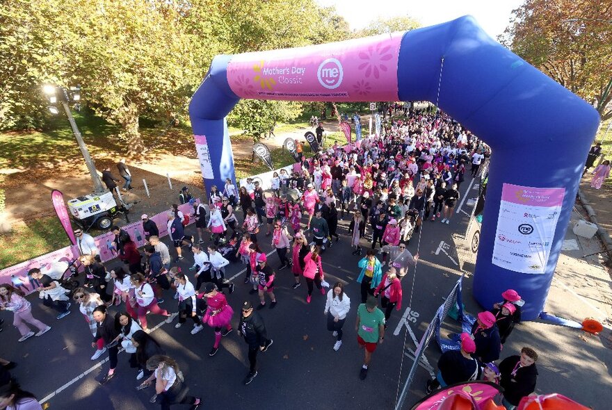 Aerial view of a group of walkers participating in the Mother's Day Classic fun run as they cross underneath a large inflatable balloon arch, trees in the background.