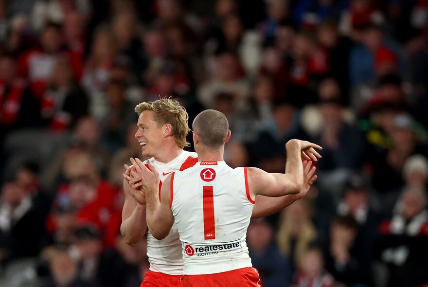 Two Sydney Swans AFL players celebrating on the field, a blurred stadium crowd behind them.