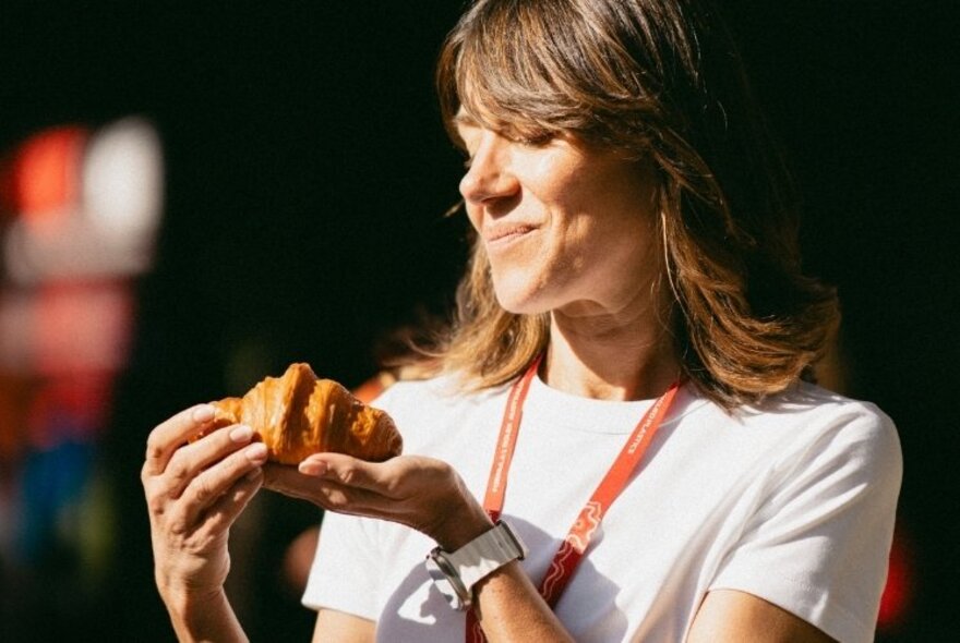Kate Reid, wearing a white t-shirt, in side profile, looking lovingly at a croissant she holds in her hand.