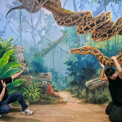 Two friends in exaggerated poses against a hand-painted interactive artwork of a jungle scene, at ArtVo.