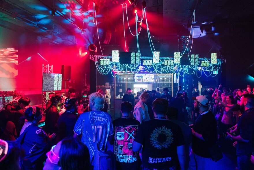 A crowded indoor event venue with red and blue stage lighting, people dancing, with screens and cables hanging from the ceiling in the back of the space.